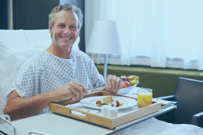 Happy man sat up in hospital bed following surgery, eating healthy lunch