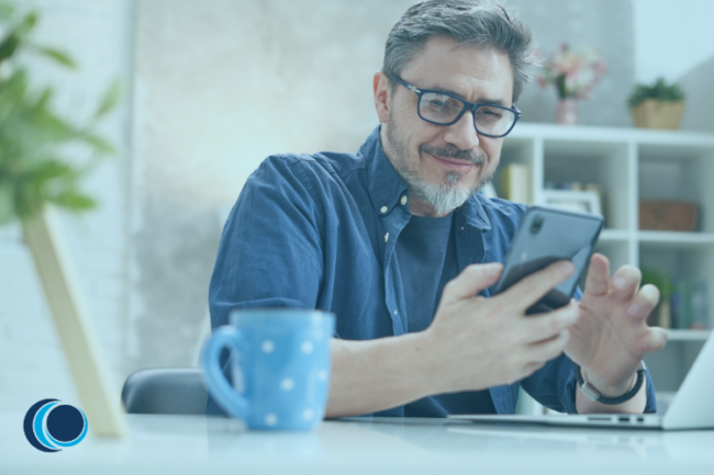 Happy middle aged man sat at desk with phone and coffee